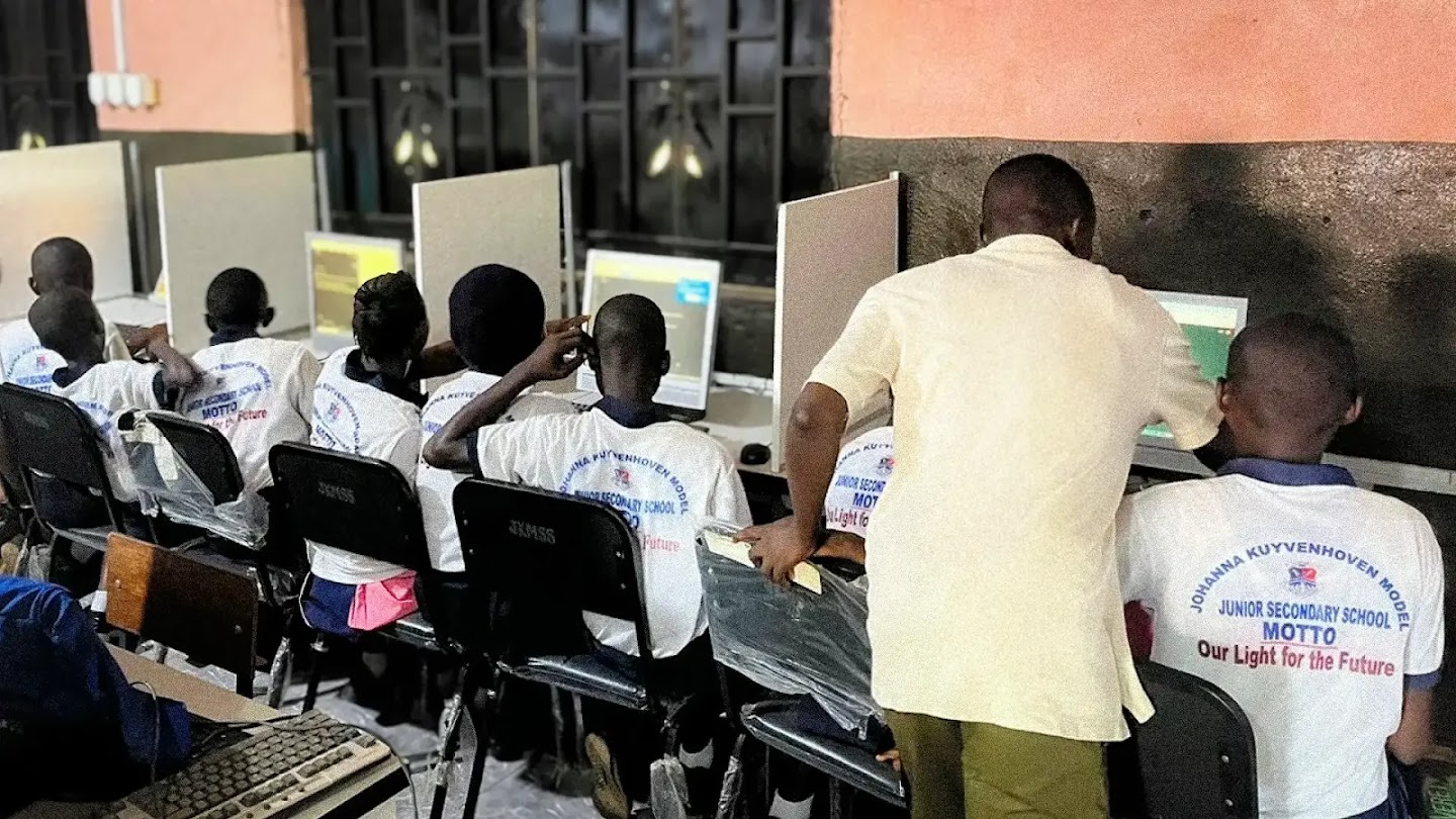 A classroom setting where several school students in matching white uniforms sit at desks with desktop computers, working on lessons. An educator stands over a student on the right, guiding them through the computer screen. The backs of the students' shirts read "Johanna Kuyvenhoven Model Junior Secondary School."